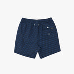 A pair of men's swim trunks featuring a dark blue and navy geometric monogram pattern, with a white snap-button waistband and a small brand patch on the side pocket