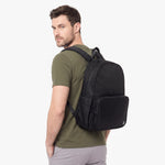 A man wearing an olive green t-shirt and gray pants is shown from the back wearing a black backpack with a front zippered pocket