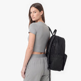 A woman wearing a gray crop top and matching sweatpants is shown from the back while carrying a black backpack on her shoulder