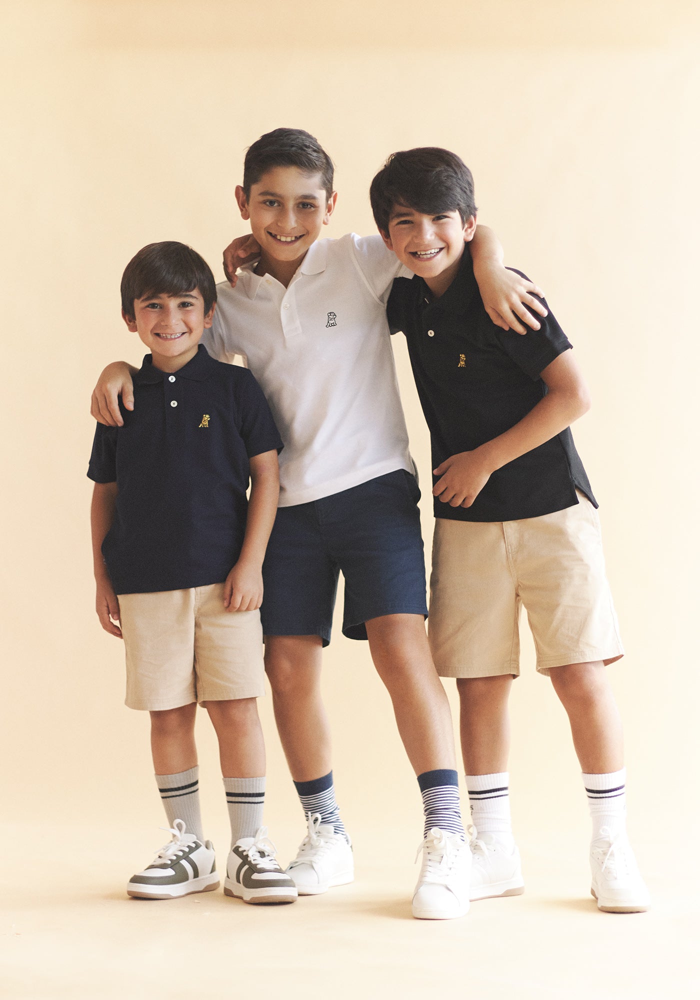 Tres niños sonrientes de diferentes edades posando juntos, vistiendo polos, shorts y zapatillas