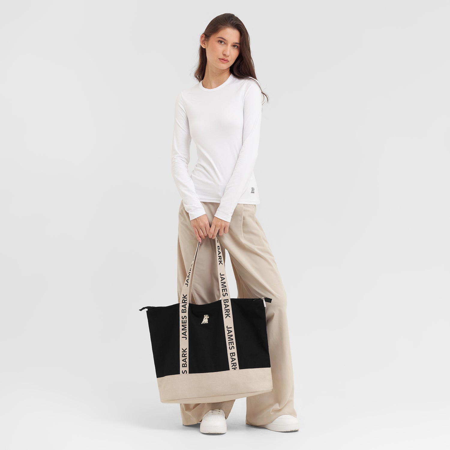 Woman holding a black and beige tote bag with 'JAMES BARK' branding on a light gray background