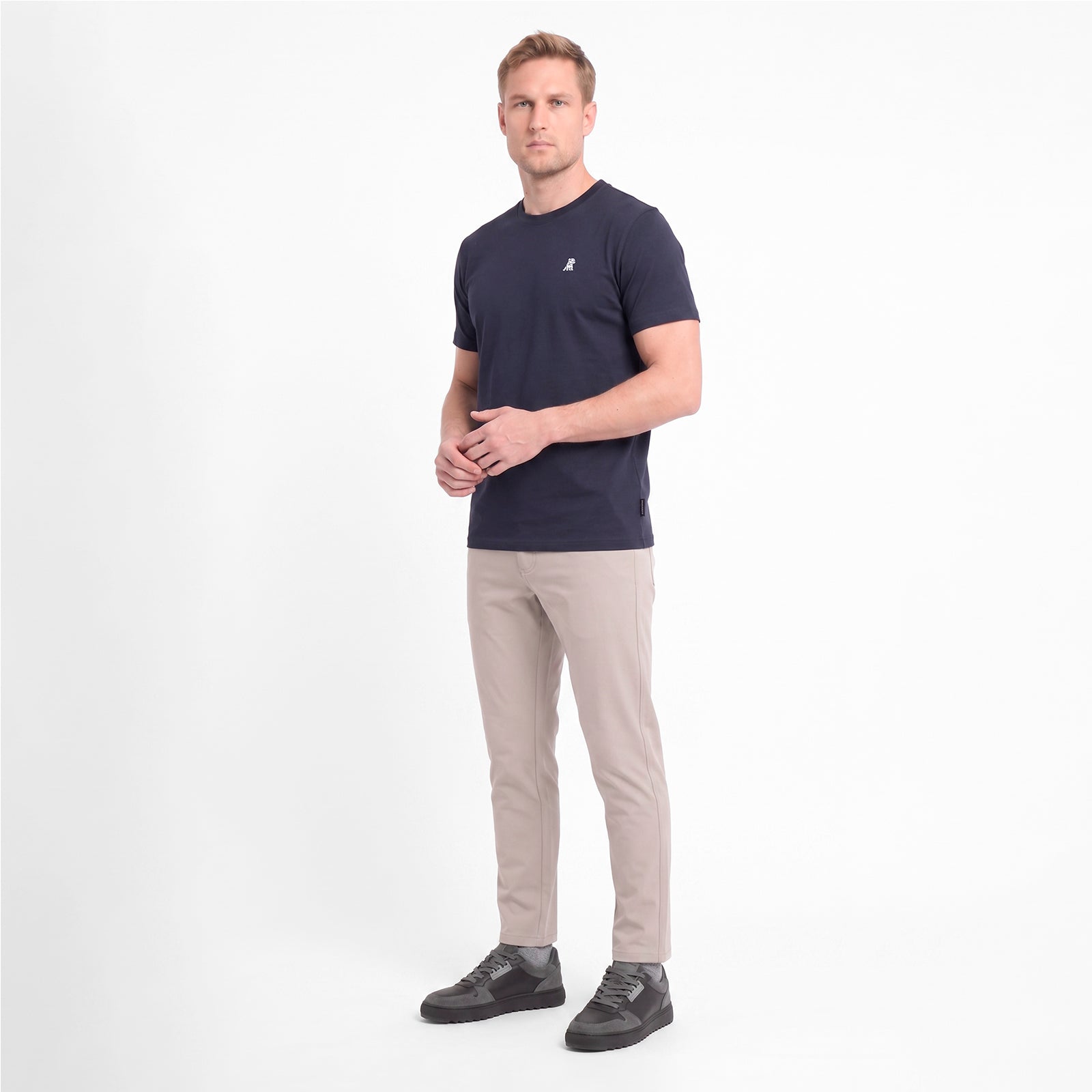 A man modeling a short-sleeve crewneck t-shirt in navy blue with a small white logo, paired with beige trousers