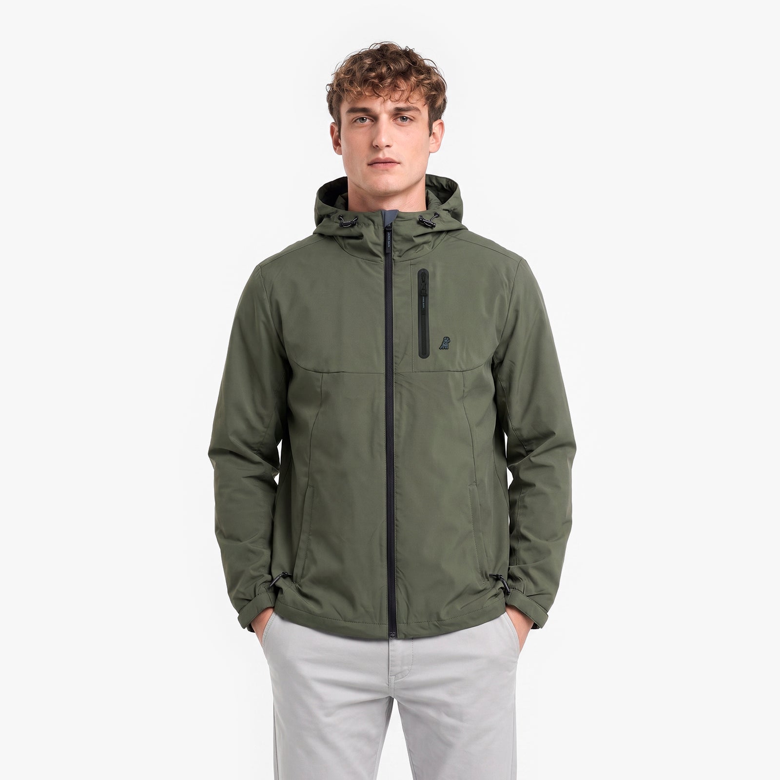 A man wearing an olive green hooded jacket with a black zipper and a small black logo on the chest, paired with light gray pants