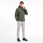 A man wearing an olive green hooded zip-up jacket, light gray trousers, and black sneakers, standing against a plain white studio background.