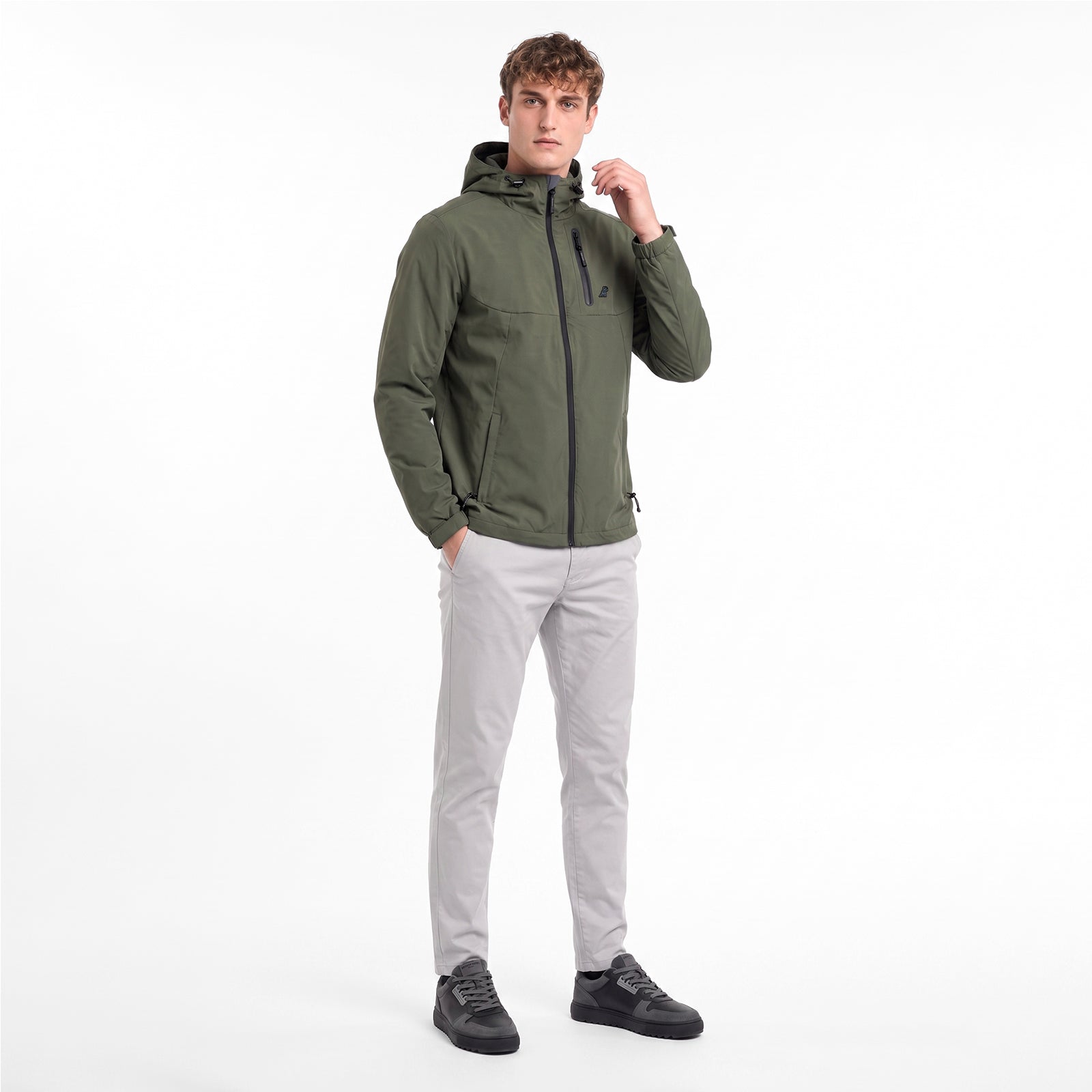 A man wearing an olive green hooded zip-up jacket, light gray trousers, and black sneakers, standing against a plain white studio background.