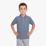 A young boy modeling a slate blue polo shirt with a small embroidered dog logo on the chest, paired with light gray pants, against a plain white background.