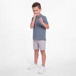 A young boy modeling a slate blue short-sleeve polo shirt, light gray shorts, and white sneakers against a clean white studio background.