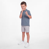 A young boy modeling a slate blue short-sleeve polo shirt, light gray shorts, and white sneakers against a clean white studio background.