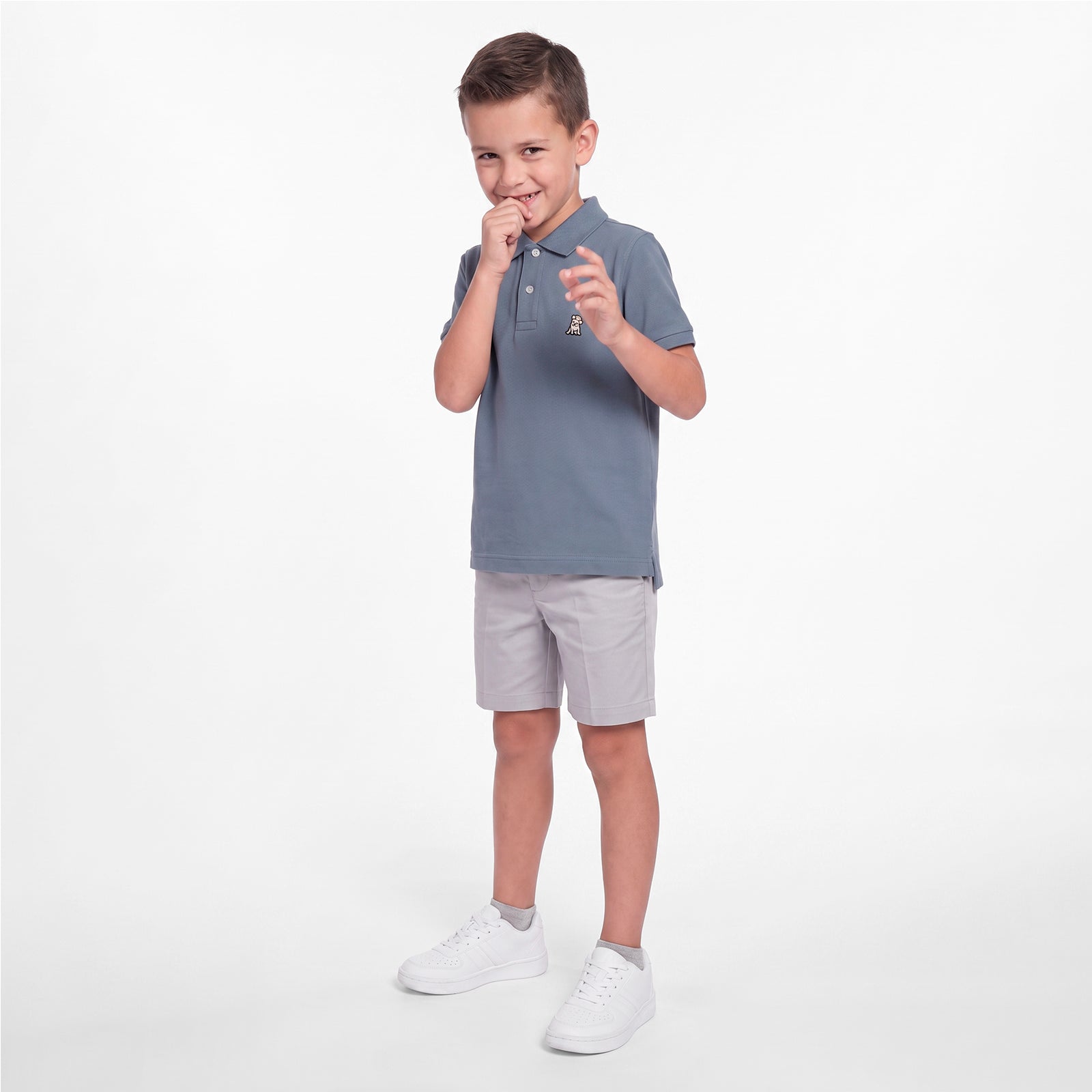 A young boy modeling a slate blue short-sleeve polo shirt, light gray shorts, and white sneakers against a clean white studio background.