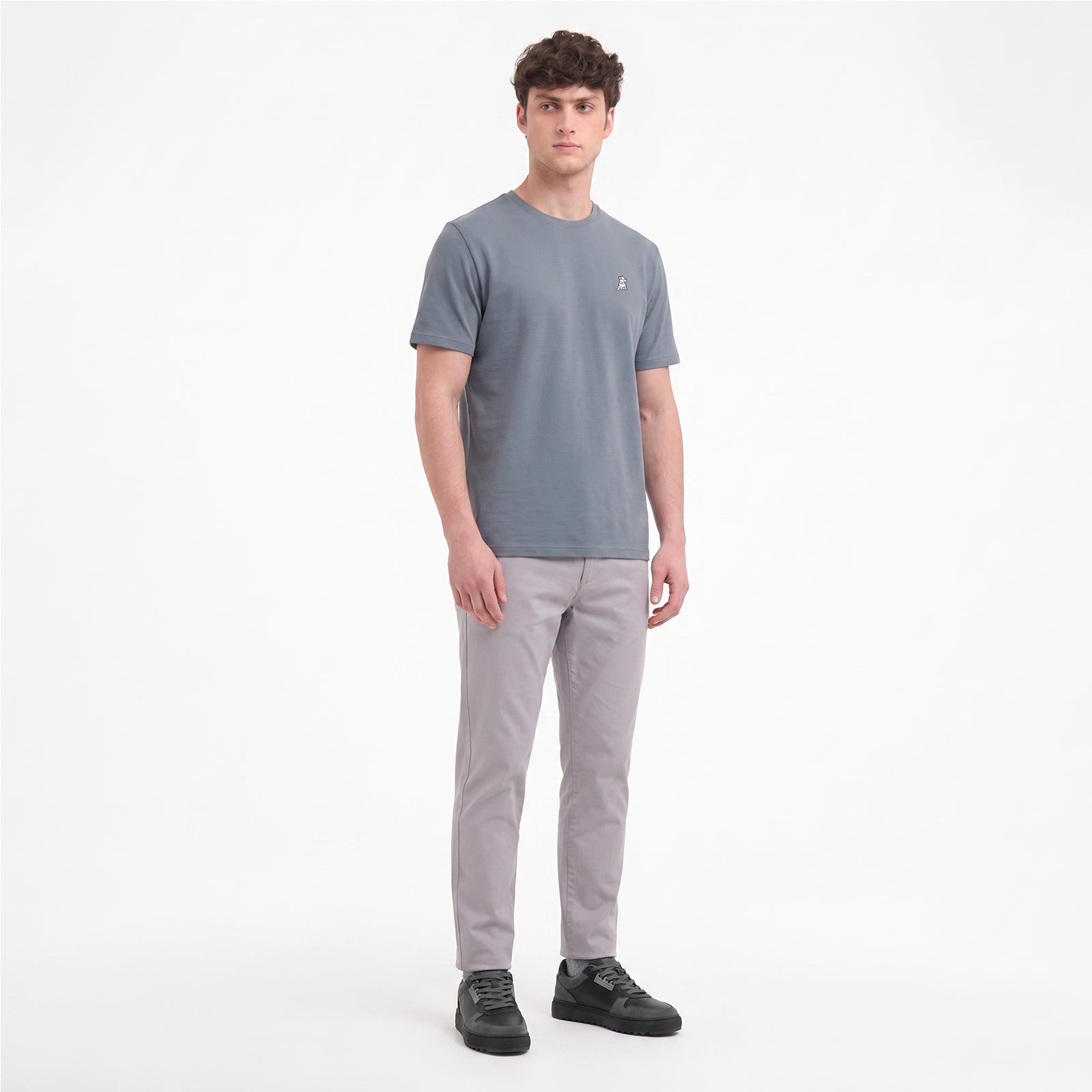 A man modeling a short-sleeve crewneck t-shirt in slate gray, paired with light gray trousers