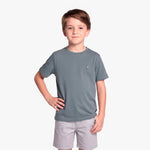 A young boy wearing a slate gray T-shirt with a small embroidered logo on the left chest, paired with light gray shorts