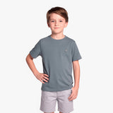 A young boy wearing a slate gray T-shirt with a small embroidered logo on the left chest, paired with light gray shorts