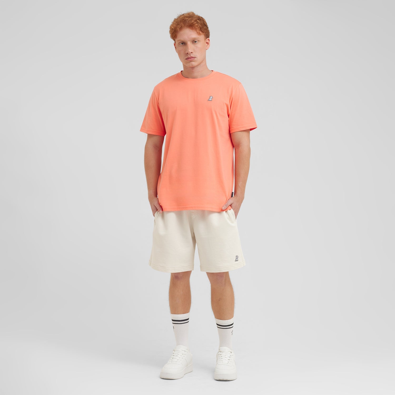 Man wearing a coral t-shirt and beige shorts on a light gray background