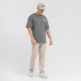 A male model wearing a gray oversized t-shirt, beige pants, a beige cap, and sneakers against a white background