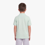 A child viewed from the back wearing a light mint green short-sleeve polo shirt and light gray pants, standing against a plain light gray studio background.