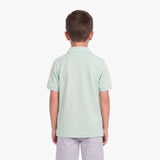 A child viewed from the back wearing a light mint green short-sleeve polo shirt and light gray pants, standing against a plain light gray studio background.