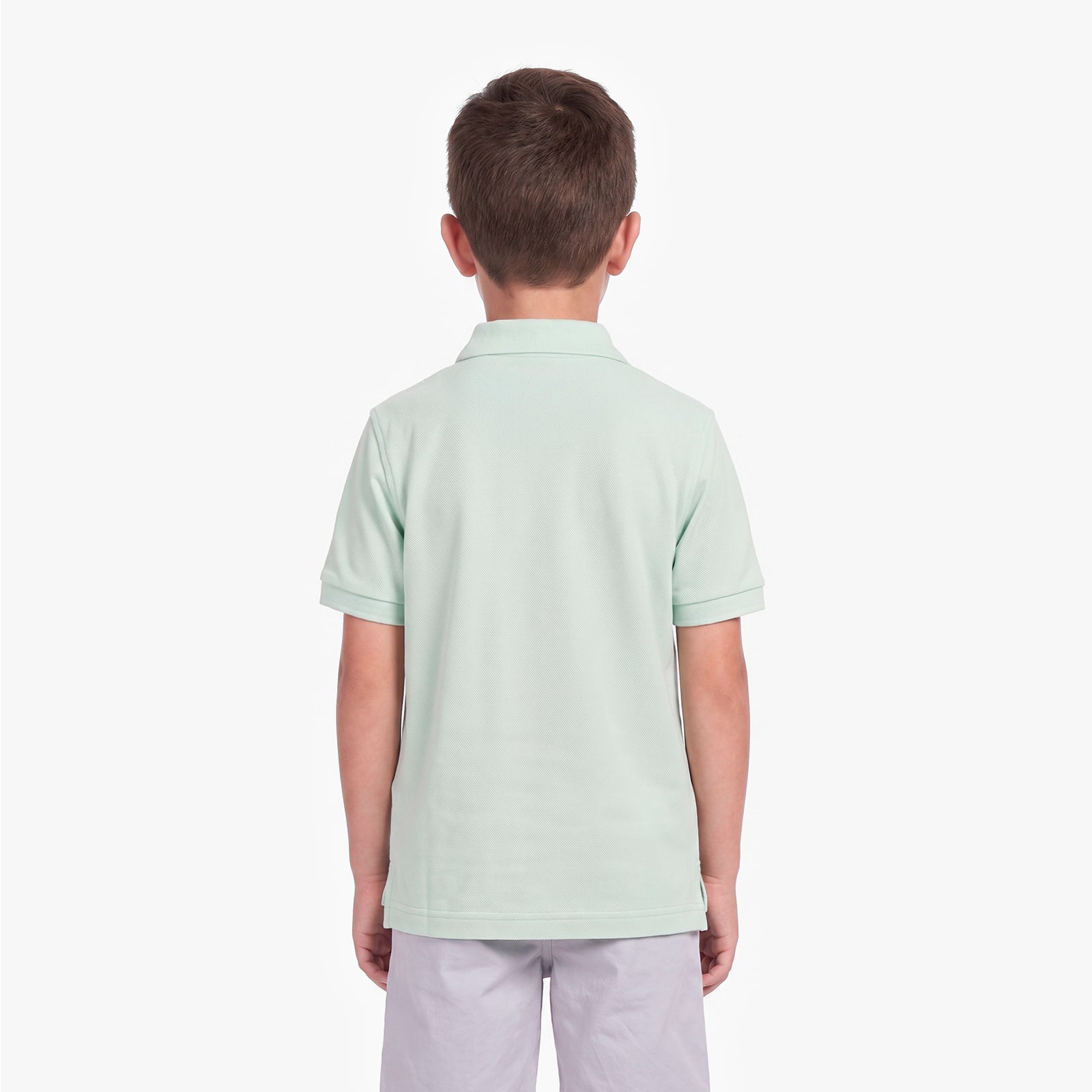 A child viewed from the back wearing a light mint green short-sleeve polo shirt and light gray pants, standing against a plain light gray studio background.