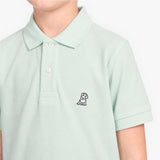 A close-up of a child wearing a pale mint green piqué polo shirt with a small embroidered dog logo on the left chest, set against a clean white background.