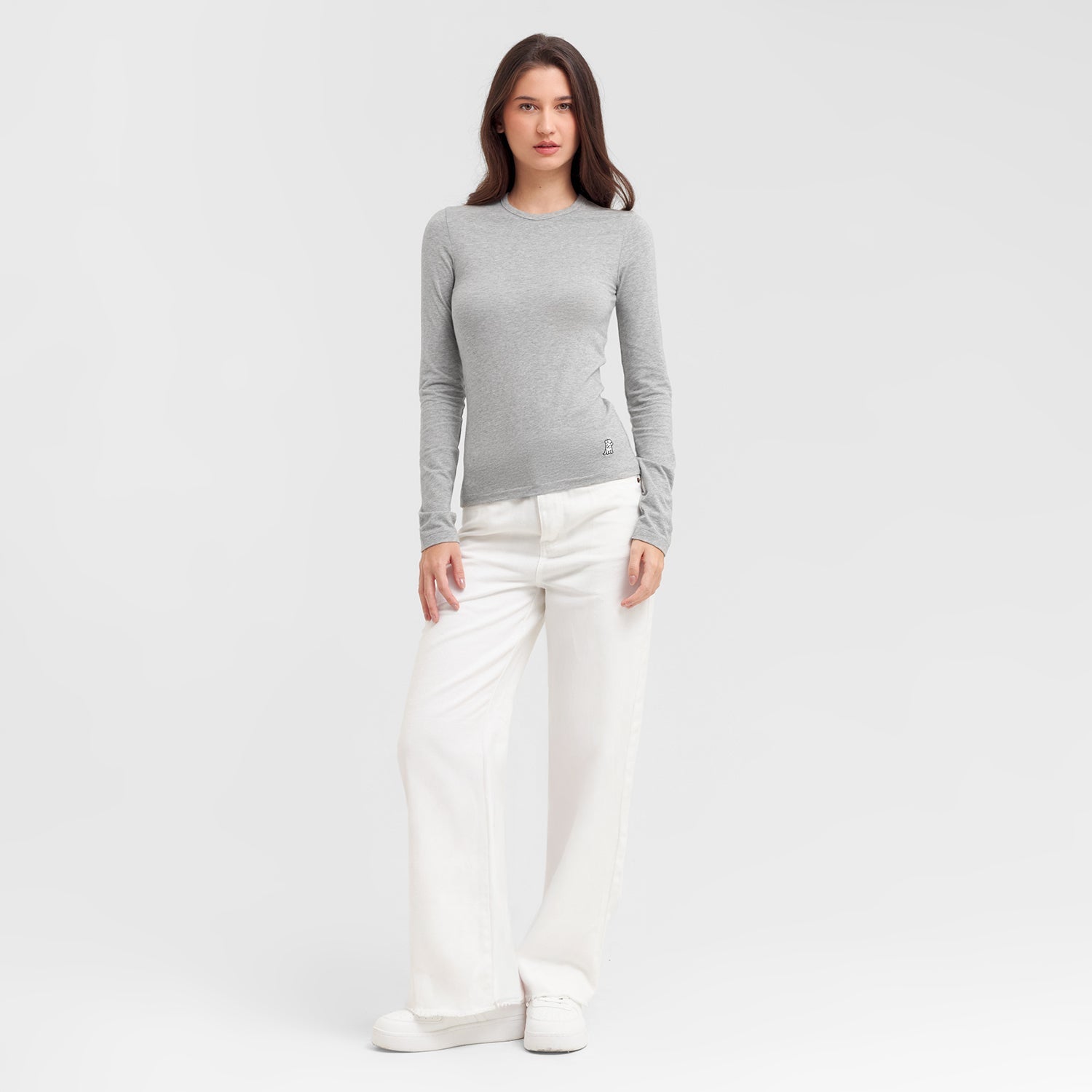 Full body view of a woman wearing a gray sweater and white pants on a light gray background