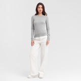 Full body view of a woman wearing a gray sweater and white pants on a light gray background