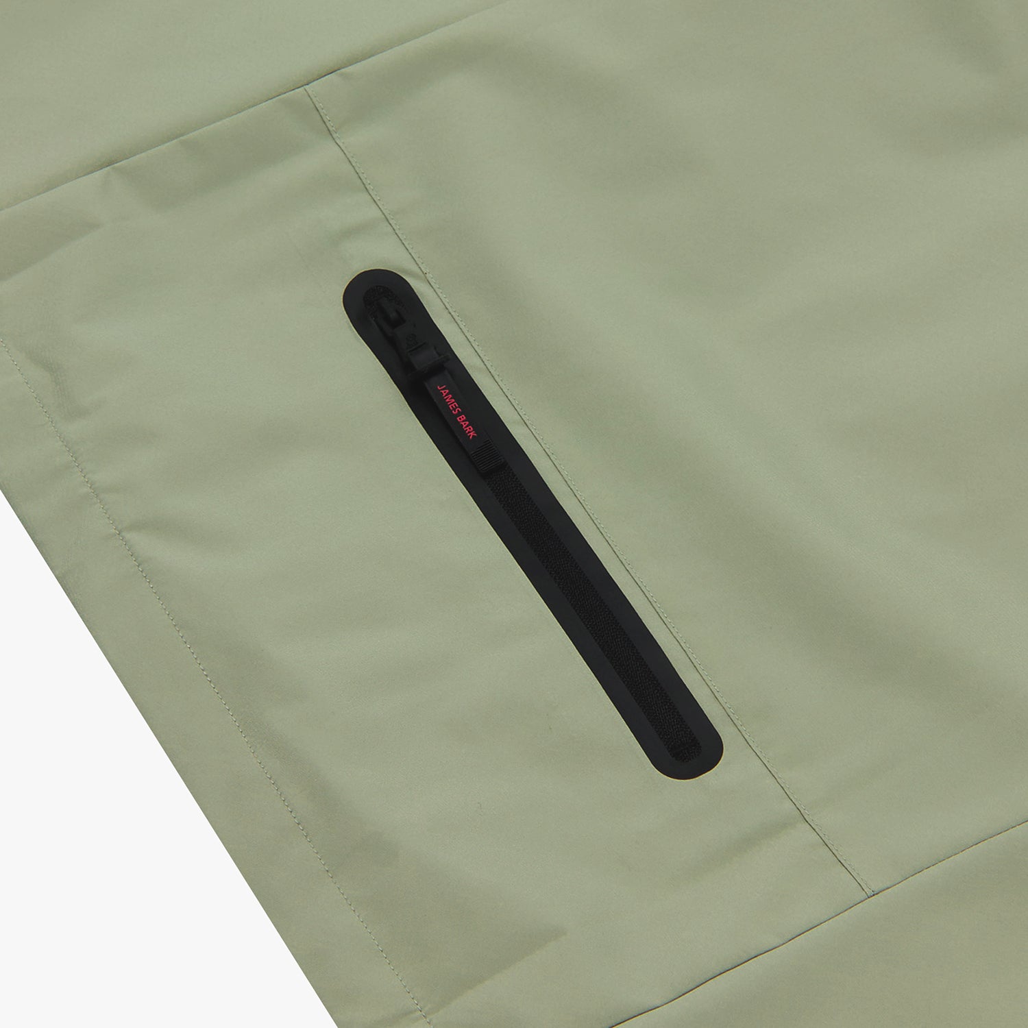 Close-up of the pocket of a sage green jacket. The black zipper pull says "JAMES BARK" in red letters.