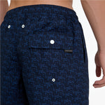 Close-up of a person wearing dark navy patterned shorts with a blue monogram print, an elastic waistband, and a flap pocket with a white snap closure. A small black brand tab reading JAMES RICE is visible on the pocket.
