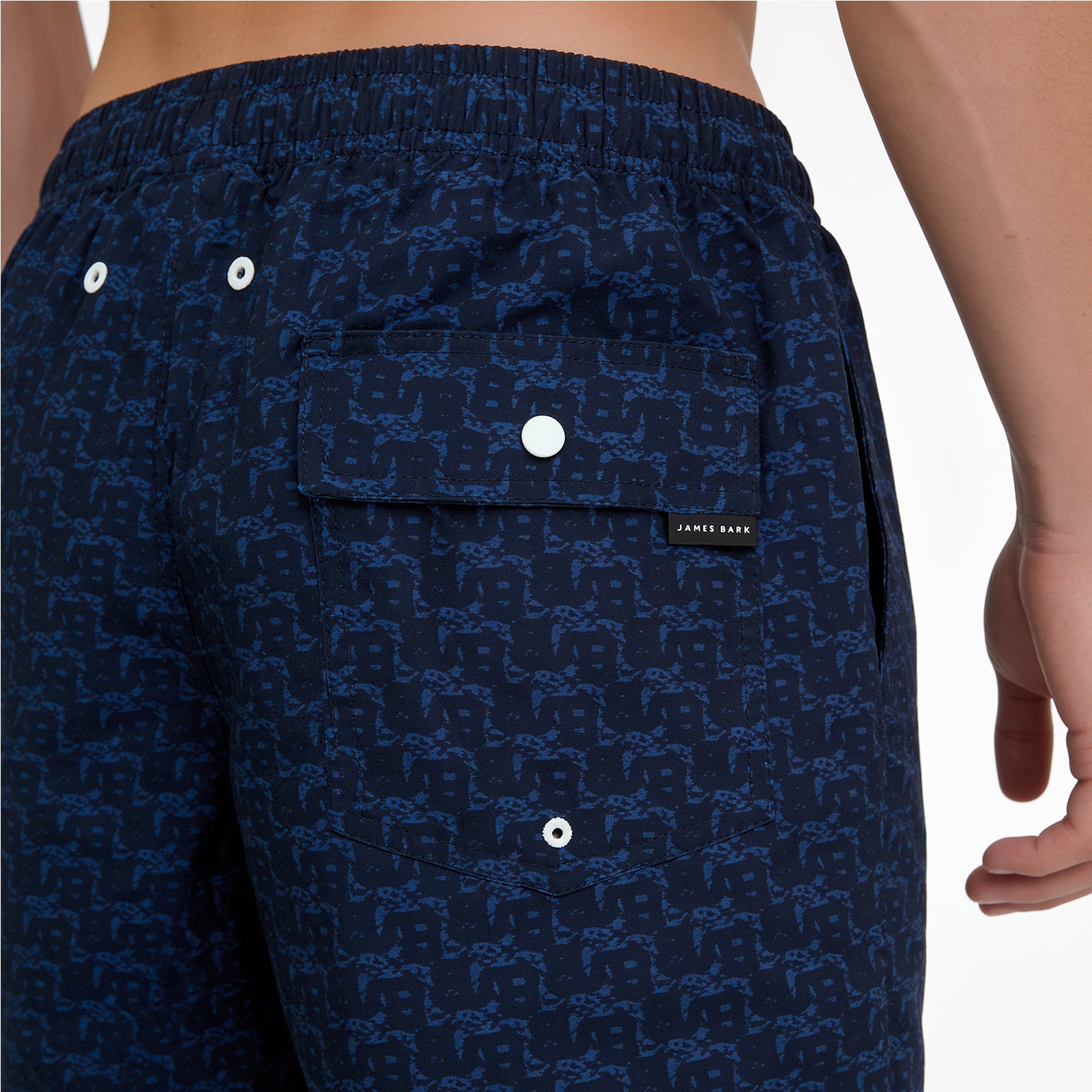 Close-up of a person wearing dark navy patterned shorts with a blue monogram print, an elastic waistband, and a flap pocket with a white snap closure. A small black brand tab reading JAMES RICE is visible on the pocket.