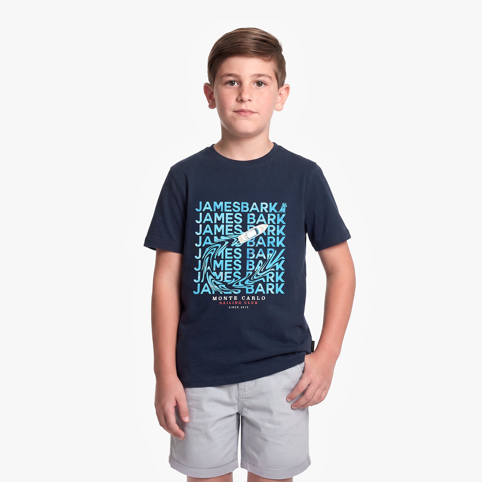 A young boy modeling a navy blue t-shirt with a light blue and white 'James Bark' graphic print on the front, paired with light gray shorts, against a plain light gray background.