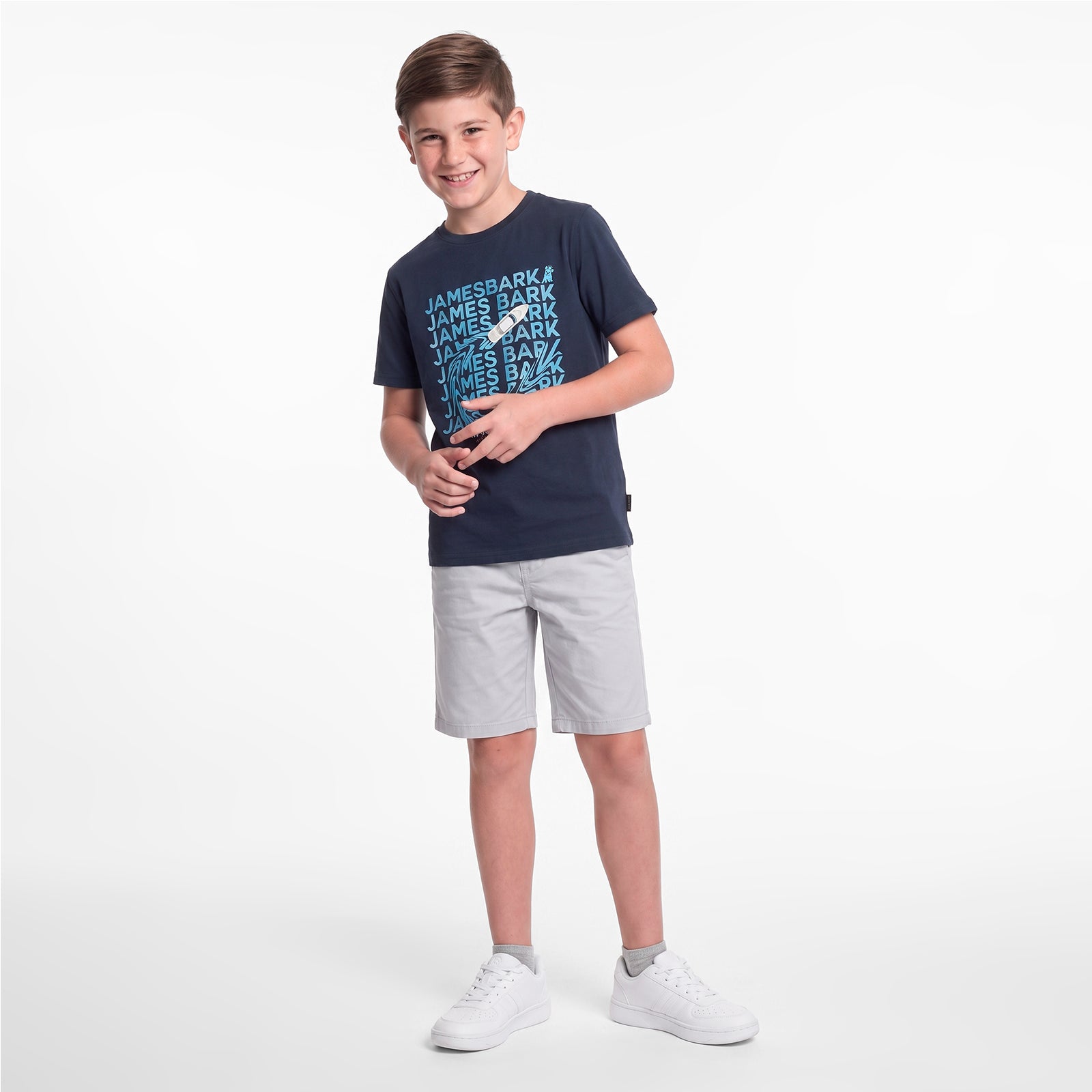 A young boy modeling a dark navy blue t-shirt with a light blue and white 'JAMES BARK' graphic, paired with light gray shorts and white sneakers
