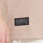Close-up of a black woven "ELEMENTS" brand tag sewn onto the hem of the beige t-shirt