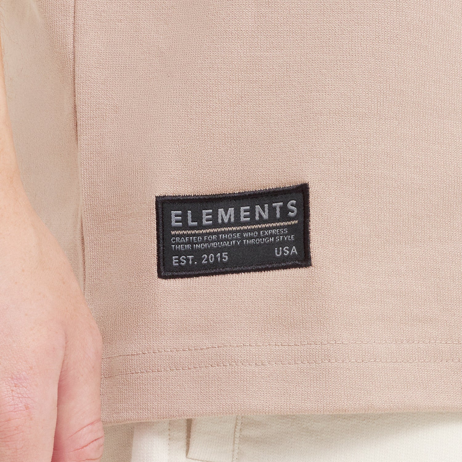 Close-up of a black woven "ELEMENTS" brand tag sewn onto the hem of the beige t-shirt