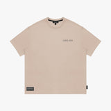 Front view of a beige t-shirt laid flat, showing a small logo on the chest and a black tag on the hem
