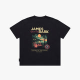 A black T-shirt shown flat against a light gray background, featuring a large graphic print on the back with the text 'JAMES BARK' and 'Luxury by the Coast'. The graphic depicts a sunset beach scene with a pool and palm trees.