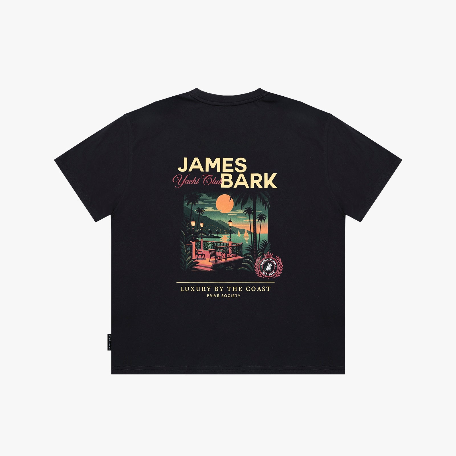 A black T-shirt shown flat against a light gray background, featuring a large graphic print on the back with the text 'JAMES BARK' and 'Luxury by the Coast'. The graphic depicts a sunset beach scene with a pool and palm trees.