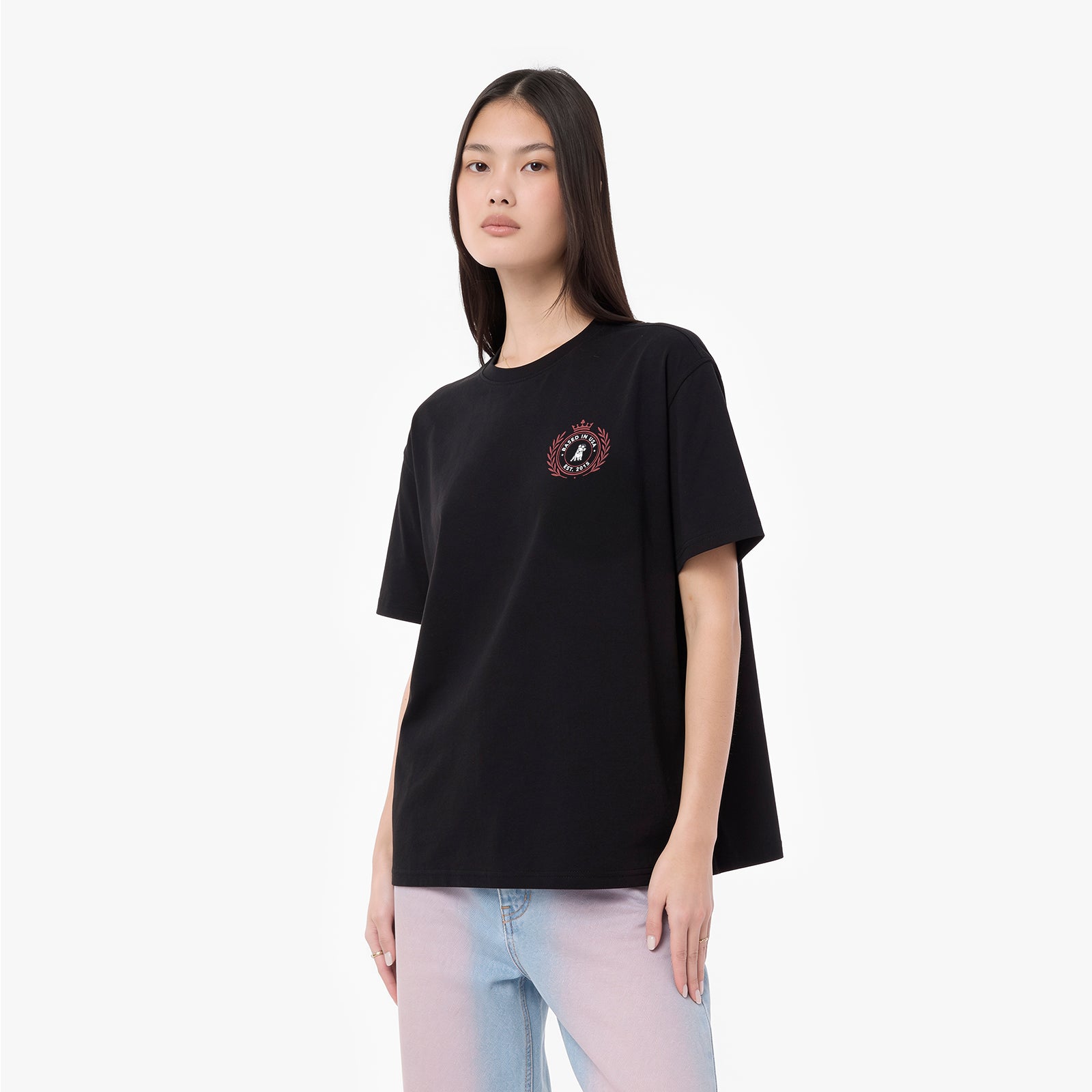 A woman modeling an oversized black short-sleeve T-shirt with a small circular graphic on the left chest, paired with light blue jeans