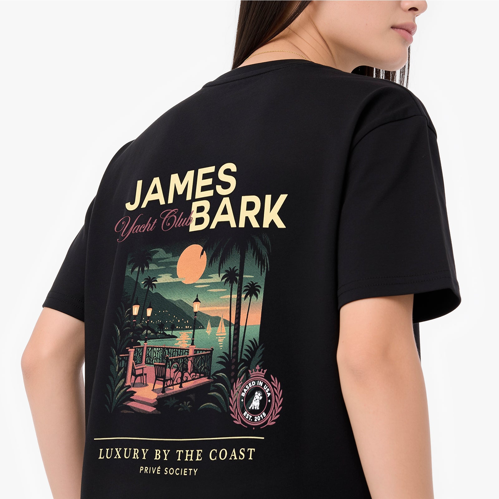 A woman wearing a black oversized T-shirt. The shirt features a large graphic print with the text 'JAMES BARK' and 'Luxury by the Coast' in cream and pink, along with a tropical beach scene illustration.