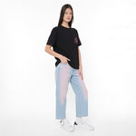 A model stands against a plain white studio background wearing a black oversized T-shirt with a small red and black chest graphic, light blue wide-leg jeans with a pink-to-blue gradient wash, white socks, and white sneakers.