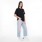 A model stands against a plain white studio background wearing a black oversized T-shirt with a small red and black chest graphic, light blue wide-leg jeans with a pink-to-blue gradient wash, white socks, and white sneakers.