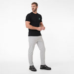 A man modeling a black T-shirt with a small green circular logo on the left chest, paired with light gray trousers and black sneakers