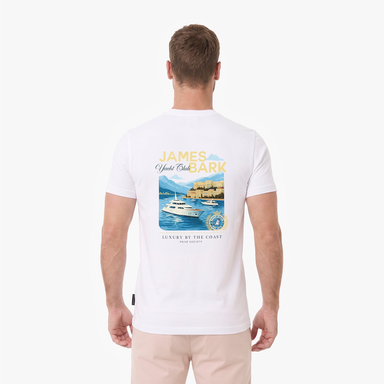 A man is viewed from behind, wearing a white T-shirt with a large graphic print on the back. The graphic features the text 'JAMES BARK' in yellow and blue, above an illustration of a yacht on blue water with mountains and a lighthouse.