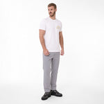 A man modeling a white T-shirt with a small circular gold and blue logo on the left chest, paired with light gray trousers and black sneakers