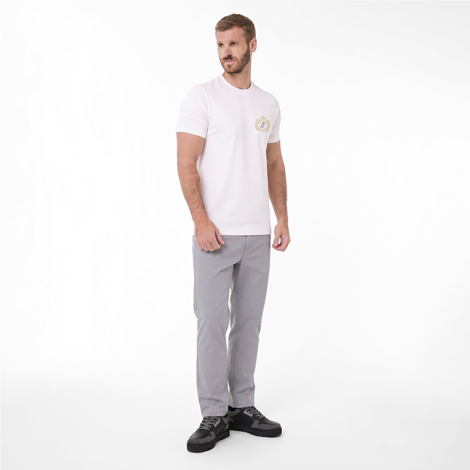 A man modeling a white T-shirt with a small circular gold and blue logo on the left chest, paired with light gray trousers and black sneakers