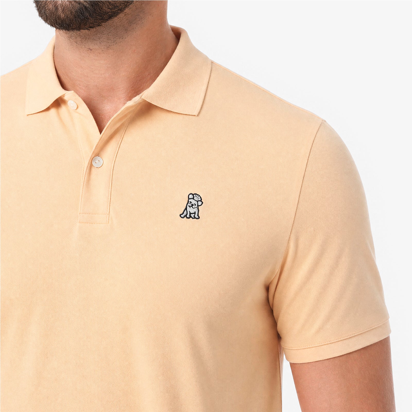 A close-up of a man wearing a light beige polo shirt with a small embroidered dog logo on the chest.