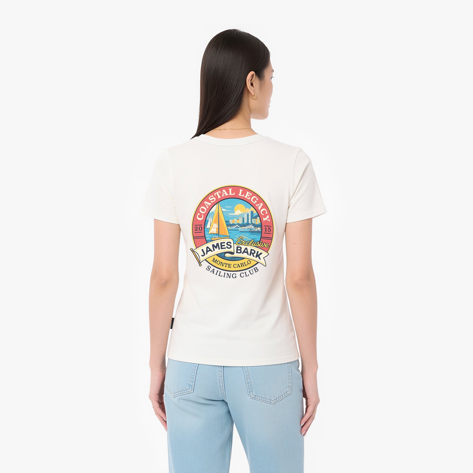 A woman viewed from the back wearing a cream-colored T-shirt with a large colorful 'James Bark Sailing Club' graphic on the back, paired with light blue jeans