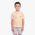 A young boy modeling a light peach-pink polo shirt with a small embroidered logo on the left chest, paired with light gray shorts, against a plain white background.