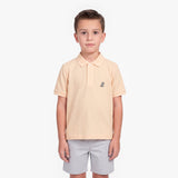 A young boy modeling a light peach-pink polo shirt with a small embroidered logo on the left chest, paired with light gray shorts, against a plain white background.