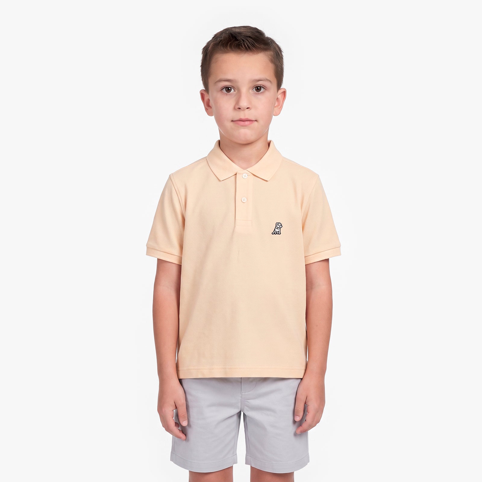 A young boy modeling a light peach-pink polo shirt with a small embroidered logo on the left chest, paired with light gray shorts, against a plain white background.