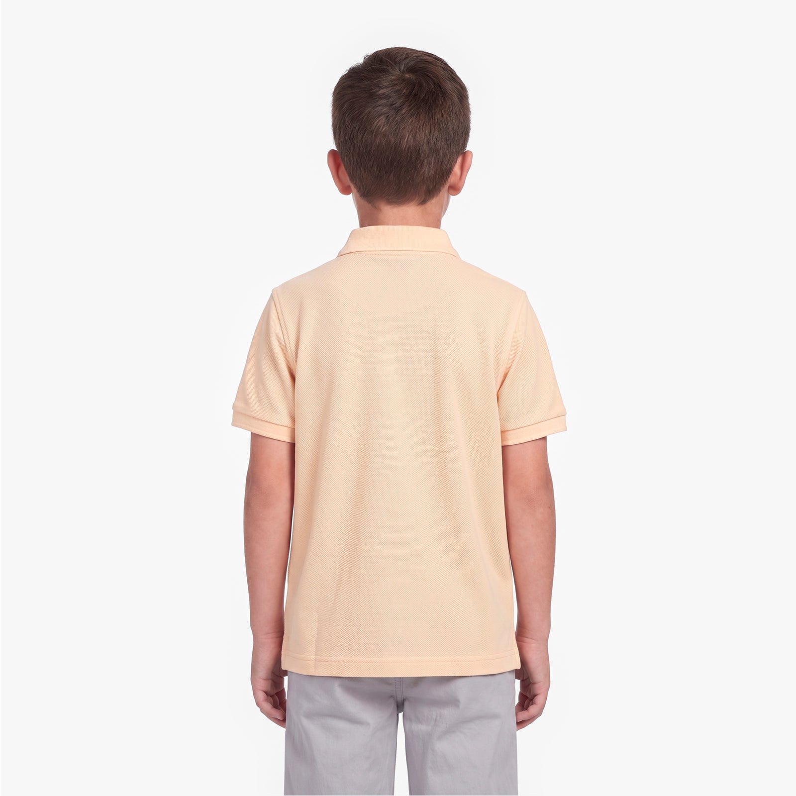 A child viewed from the back wearing a short-sleeve polo shirt in a pale peach-beige color, paired with light gray pants, against a plain white background.
