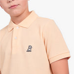 A close-up of a child wearing a light peach-pink piqué polo shirt with a small embroidered dog logo on the chest, set against a plain white background.