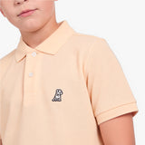 A close-up of a child wearing a light peach-pink piqué polo shirt with a small embroidered dog logo on the chest, set against a plain white background.